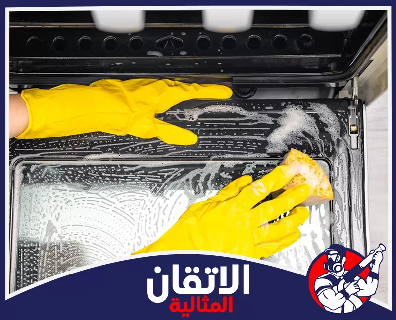 صيانة افران كهربائية بالظهران 0567008659 شركة الإتقان المثالية تنظيف باب فرن كهربائي باستخدام إسفنجة ورغوة منظف، مع ارتداء قفازات صفراء لإزالة الدهون والبقع الصعبة